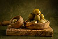 A wood bowl of green stuffed olives with olive oil pouring on top and sitting on wooden cutting board